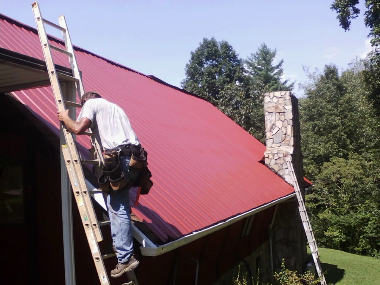 Roofer on a ladder beside a red metal roof and stone chimney — Soffit & Fascia Repair by Roofing Brothers in Meherrin